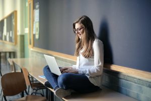 Woman sitting on a table and typing on a computer next to a blackboard