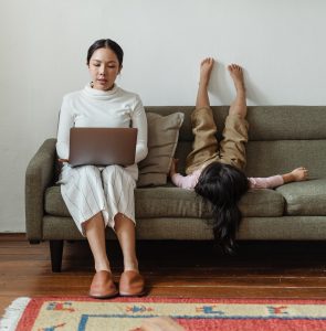 A woman is working on her computer, and her daughter is upside down on a couch