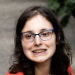 Young caucasian women with shoulder length brown hair. Smiling. Wearing glasses and an orange blouse