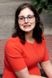 Young caucasian woman with shoulder length brown hair wearing glasses and an orange blouse.
