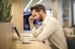 View from the side. Man sitting at table looking at open computer with coffee cup sitting next to him. Hand at forehead. Appears to be thinking. Wearing white shirt, short brown hair.