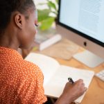 A woman is writing at her desk with a pen and a notebook in front of a computer