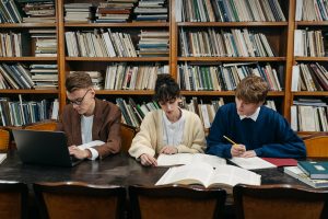 Three people are sitting in front of bookcases in a library while writing on a computer or in paper notebooks